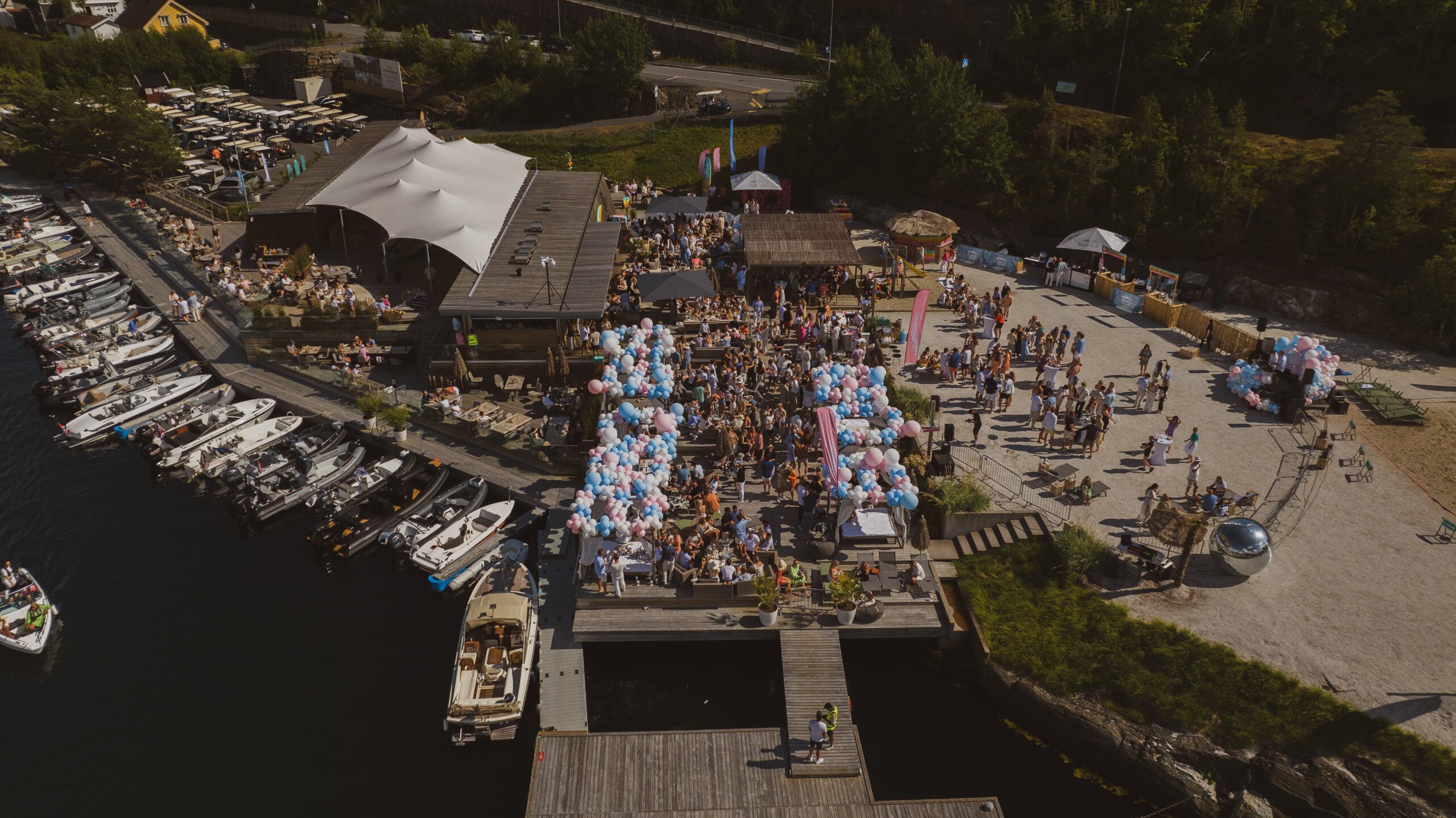 Aerial view of a lakeside festival with boats docked along the left, a large white tent, food stalls, and a crowd gathered on the dock and sandy area.
